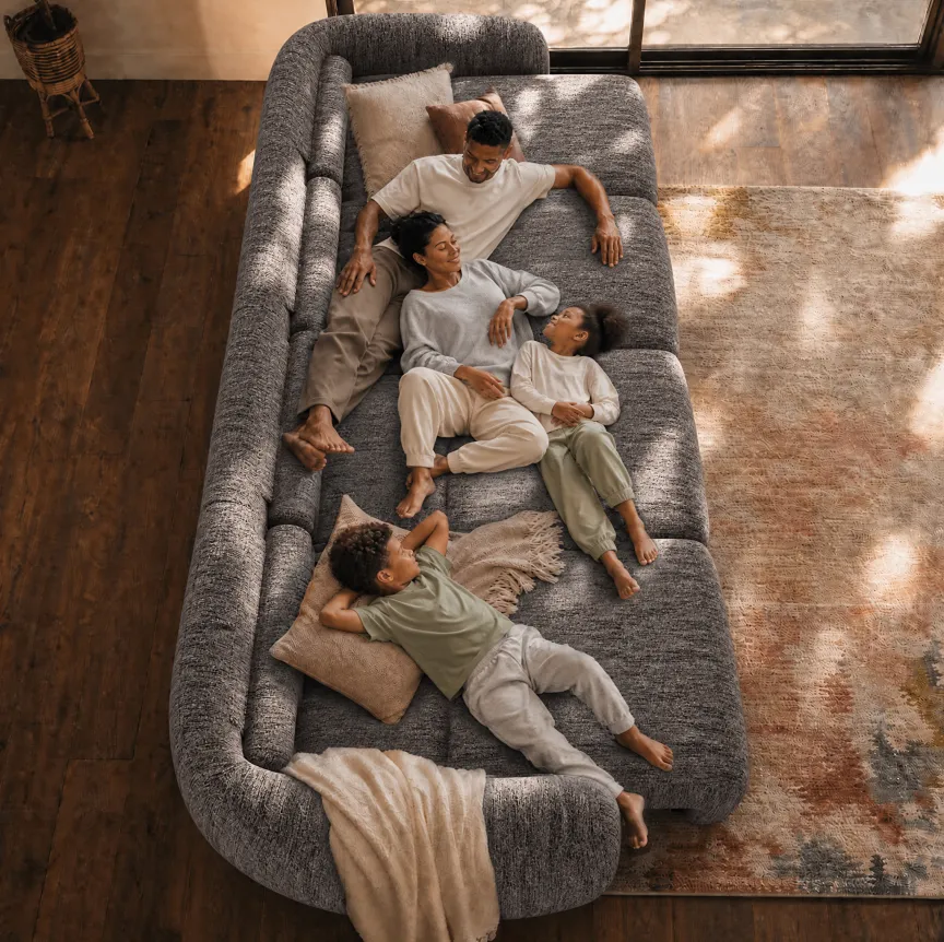 Overhead view of a family of four relaxing together on a spacious gray sofa bed in a sunlit living room.
