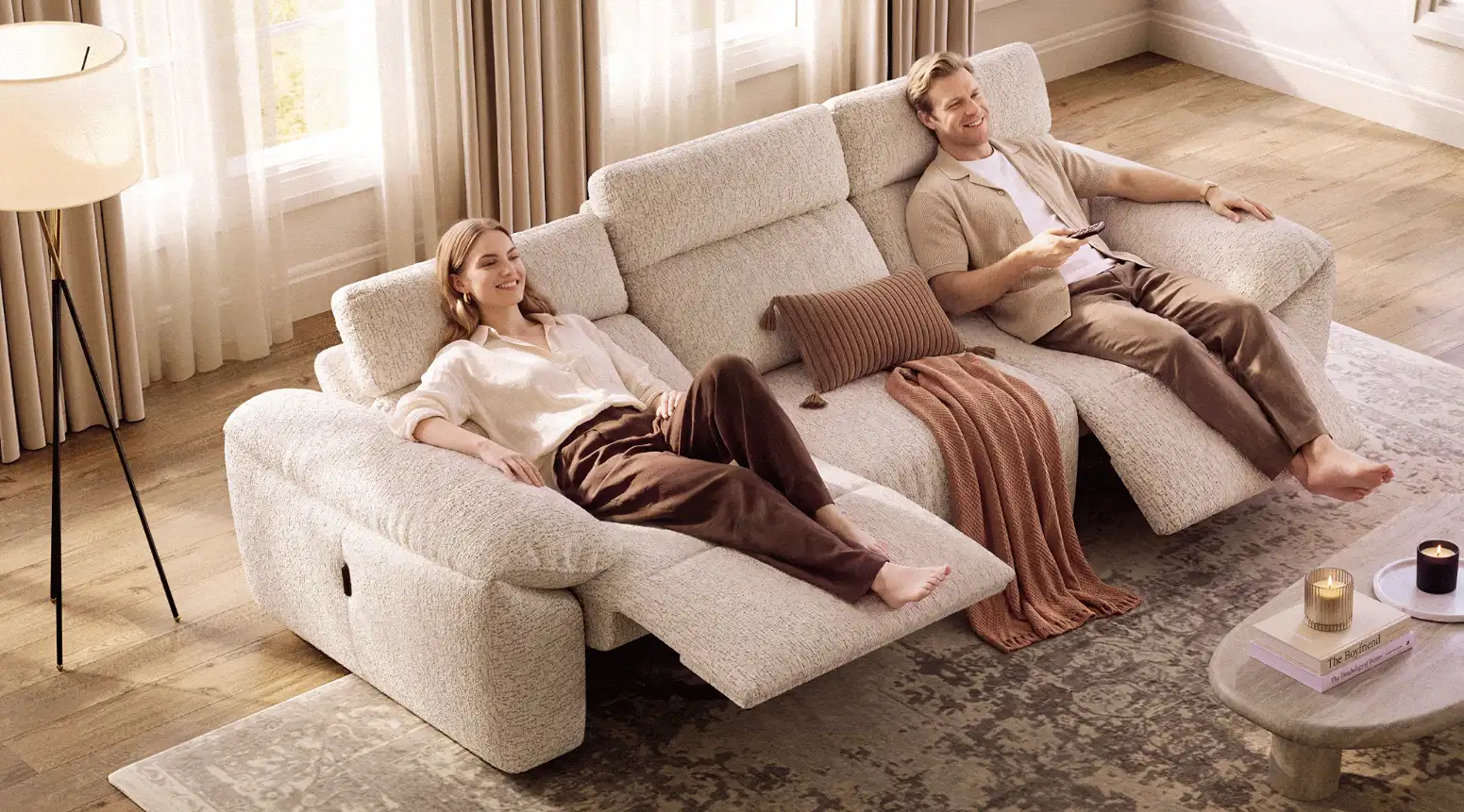 Two people reclining on a beige power sofa with extended footrests and adjustable headrests in a bright living room.