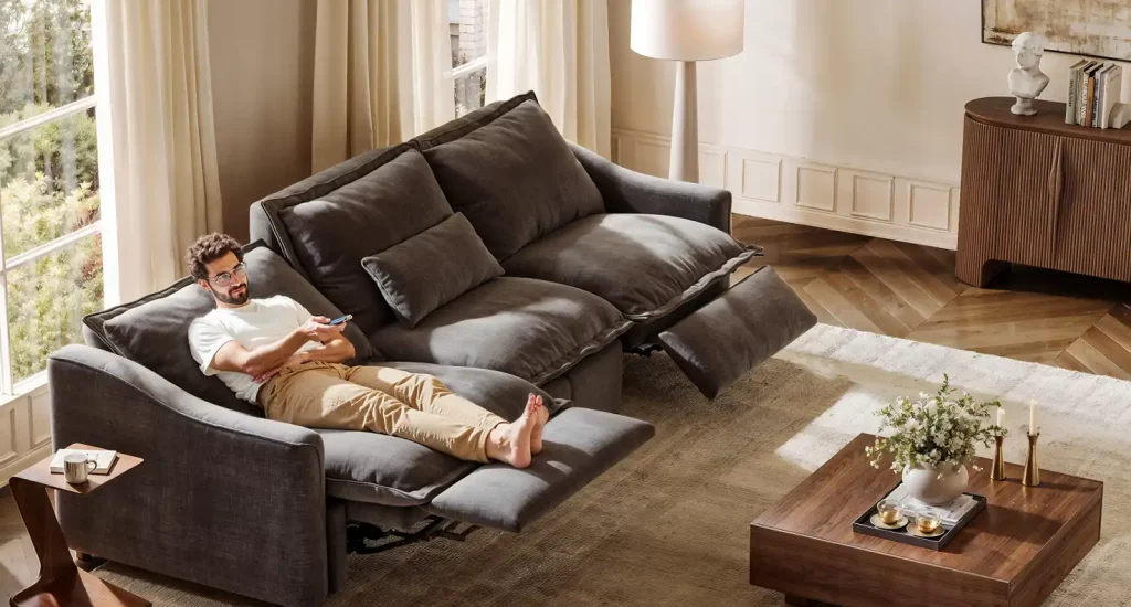 Man relaxing on a deep-seat gray sofa with extended footrest in a bright modern living room.