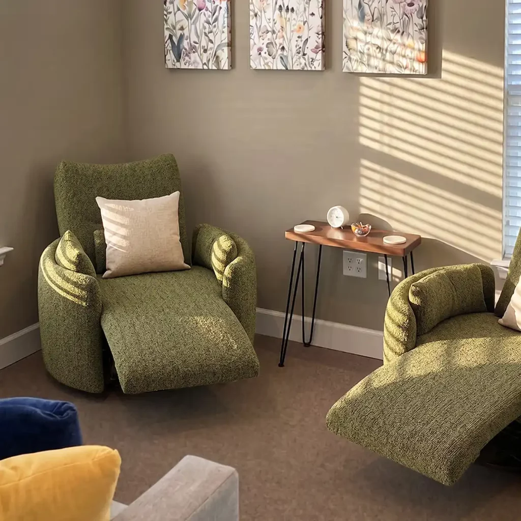 Two green upholstered reclining chairs with a small wooden side table in a sunlit living room corner.