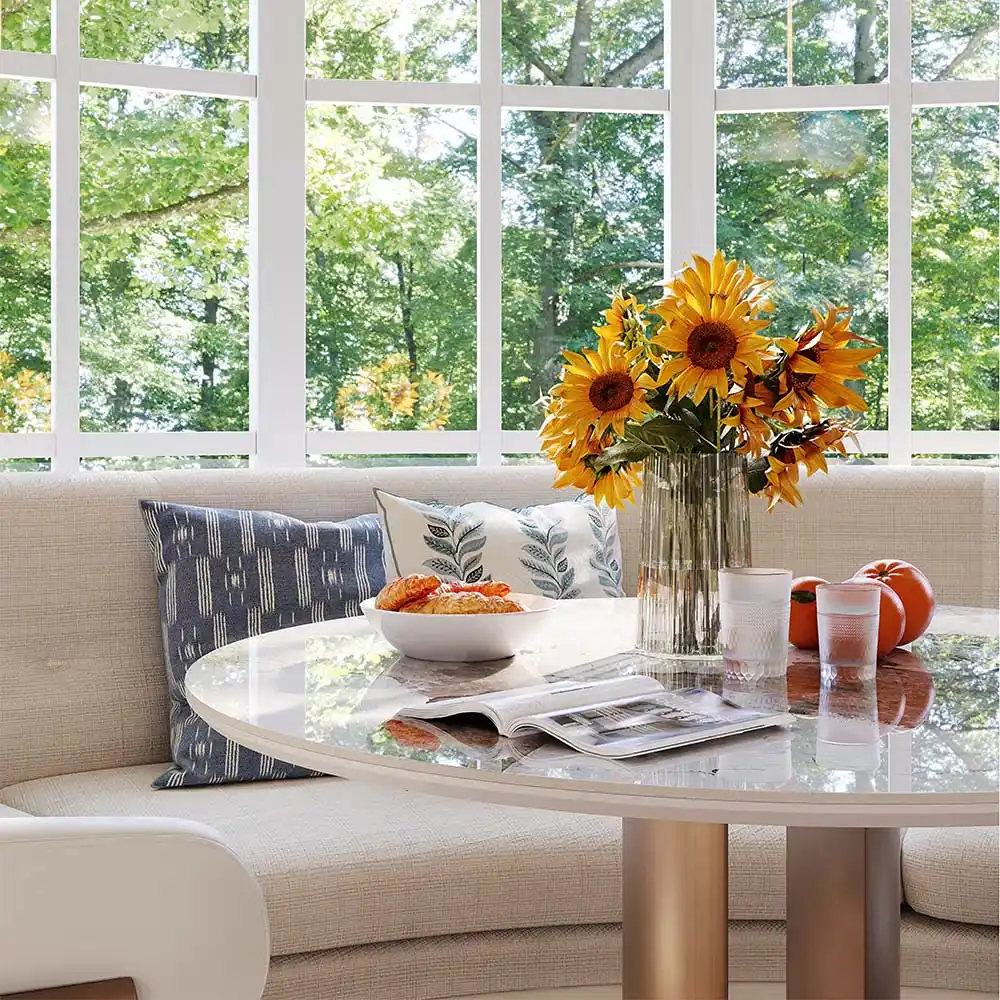 Bright dining nook with a curved upholstered banquette, round table, and sunflowers by large windows.