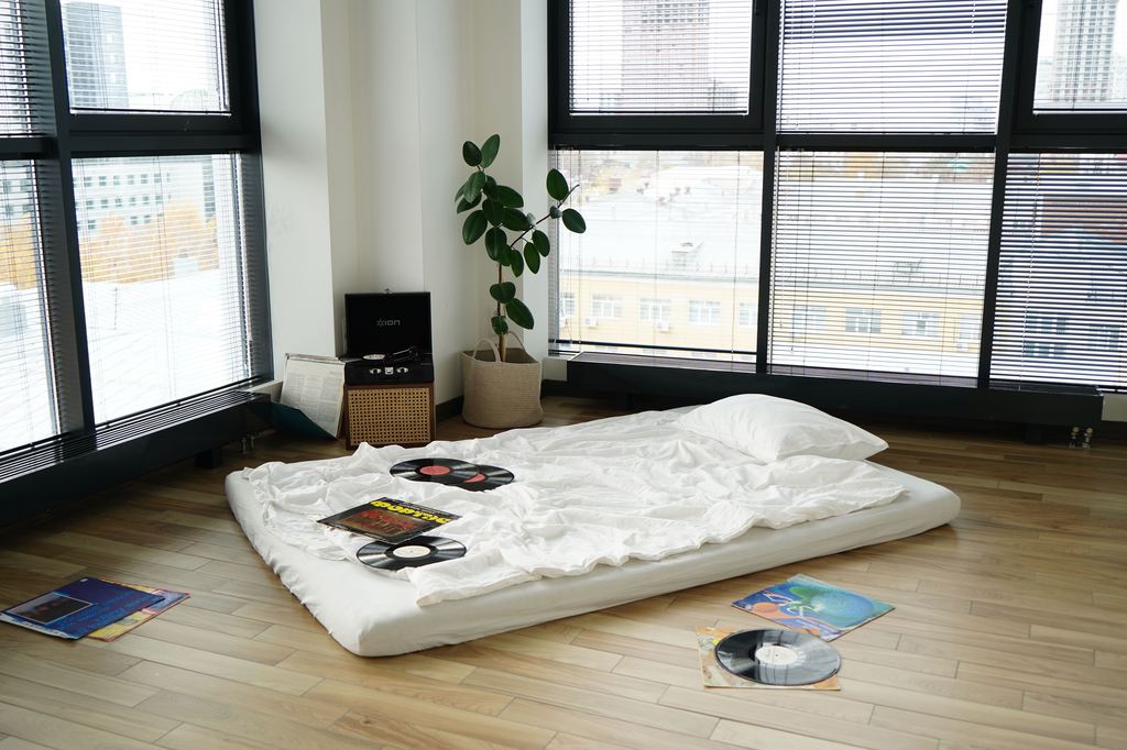 Low floor mattress with white bedding in a bright studio apartment with large windows and wooden flooring, offering a simple guest sleeping setup.