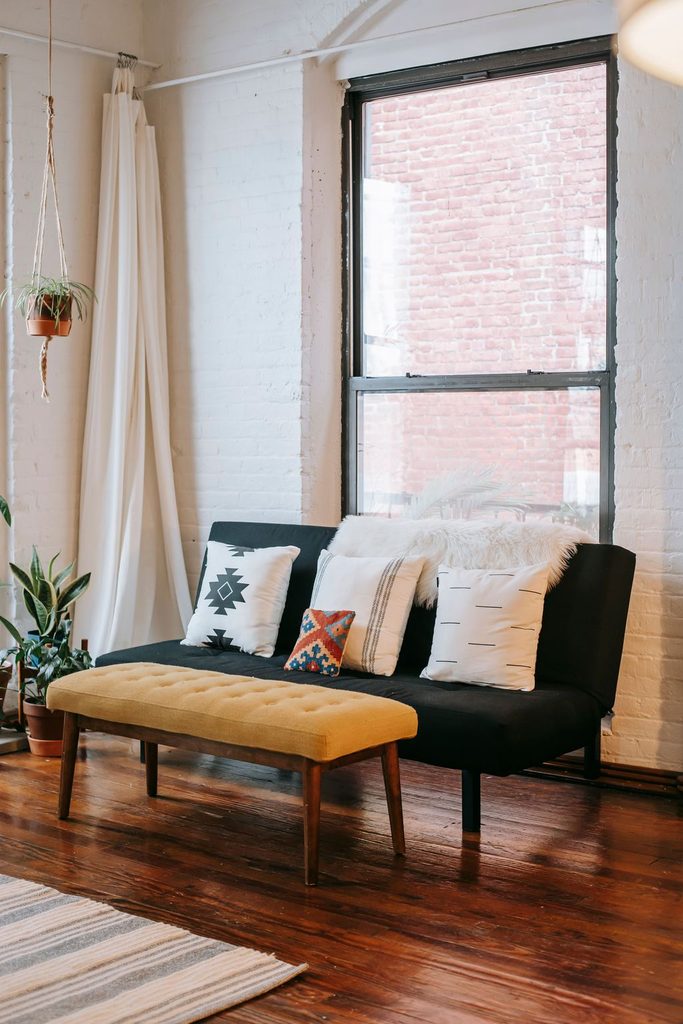 Black futon-style sofa with decorative pillows and a mustard yellow bench in a cozy, small living room.