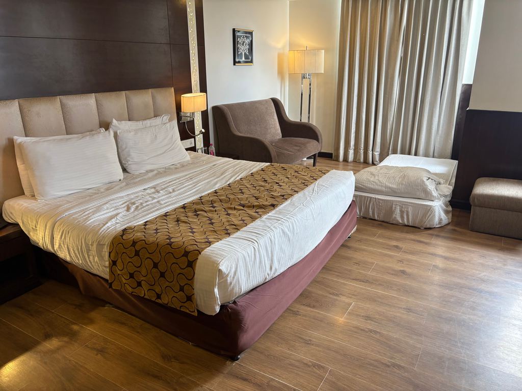 Spacious hotel-style bedroom with a large bed, armchair, floor lamp, and a separate sleeping area near the window.