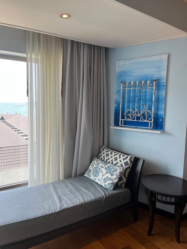 Gray chaise lounge with blue and white patterned pillows beside a round dark wood table in a room with light blue walls and ocean views.
