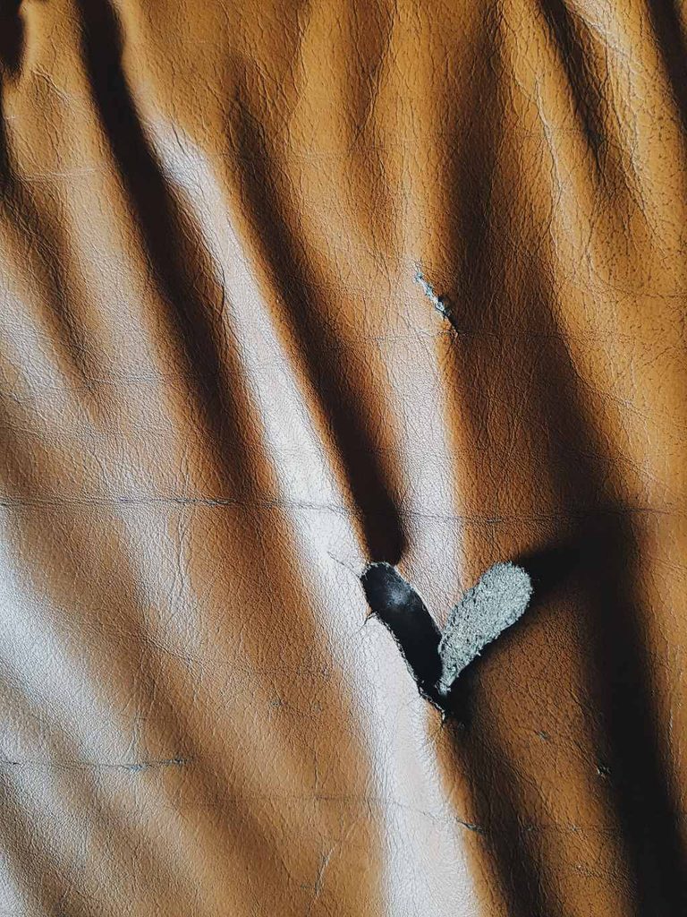 Close-up of brown leather upholstery with a tear exposing the lighter backing underneath, surrounded by creased and worn surface areas.