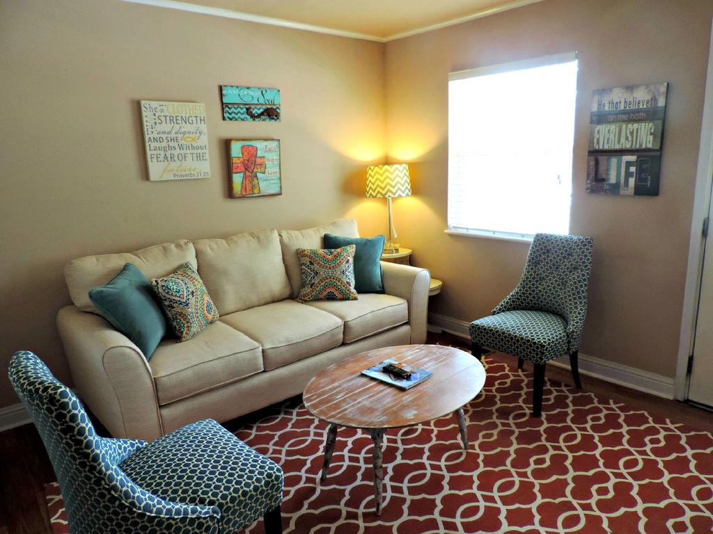Small den furniture layout with a beige three-seat sofa, two blue patterned accent chairs, an oval wooden coffee table, and a red patterned rug.