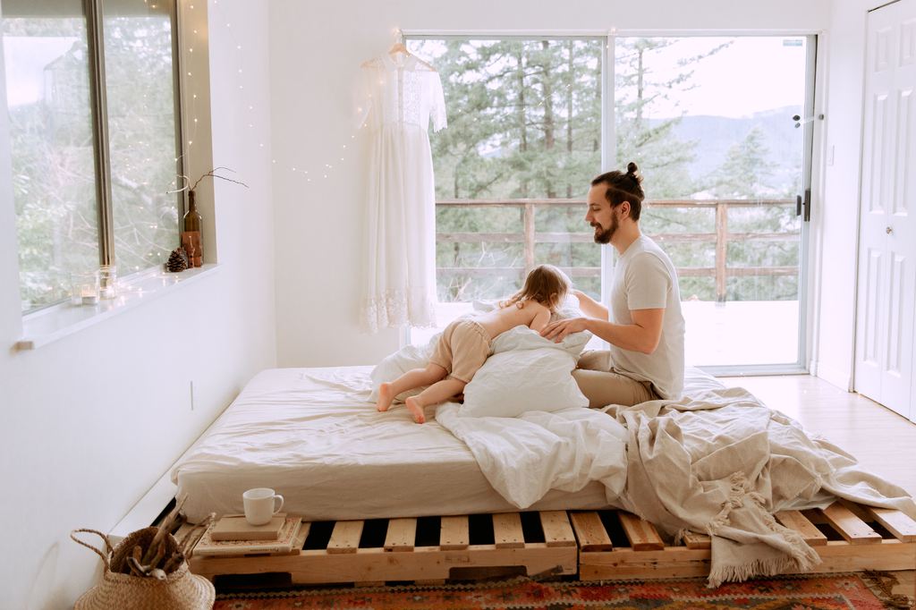 Man and child sitting on a mattress atop a DIY wooden pallet bed frame in a bright, airy room.