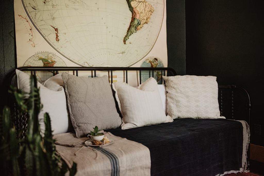 Black metal daybed with neutral throw pillows and a dark blanket beneath a vintage world map in a cozy guest room.