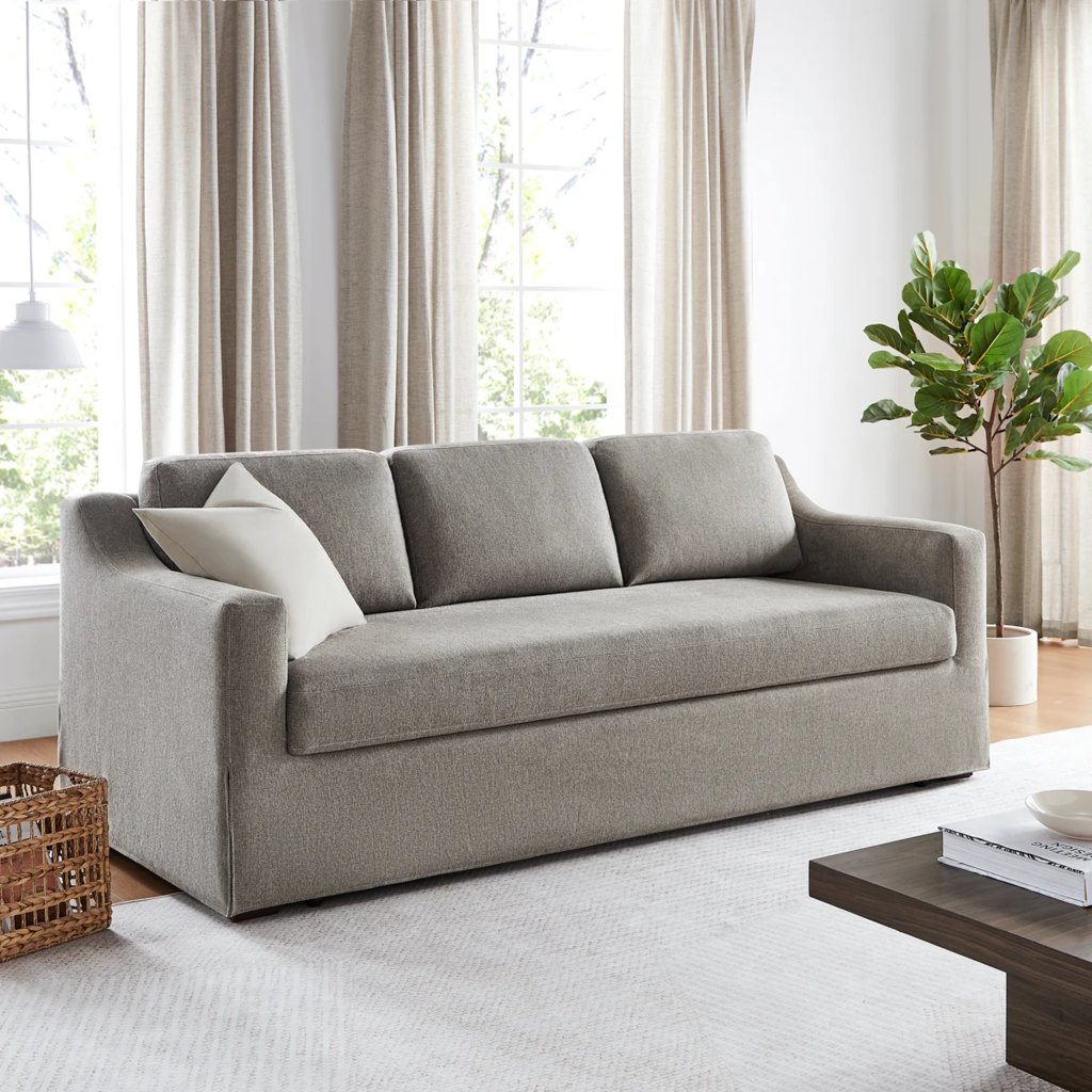 Gray three-seater sofa bed with three back cushions and white throw pillows in a bright living room with tall windows and neutral decor.