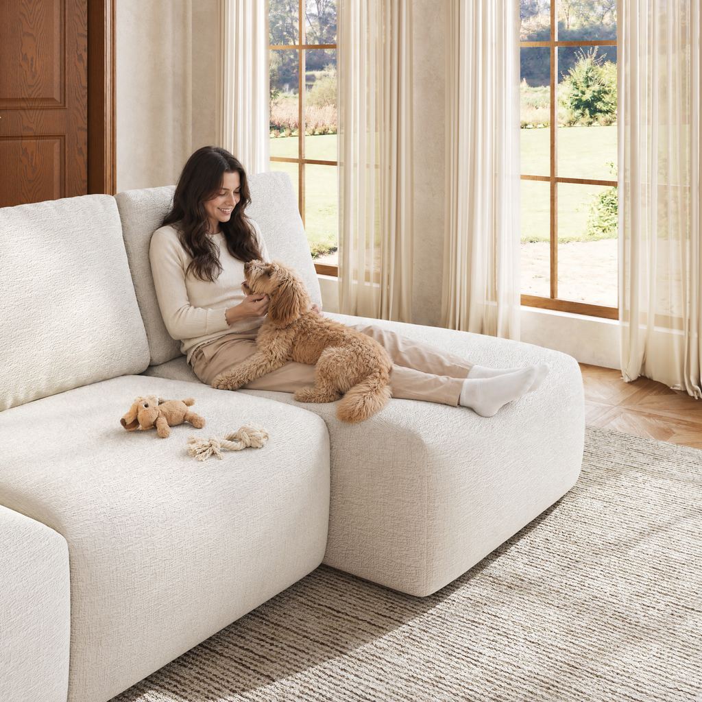 Person relaxing with a small dog on a white sectional sofa in a bright living room with large windows.