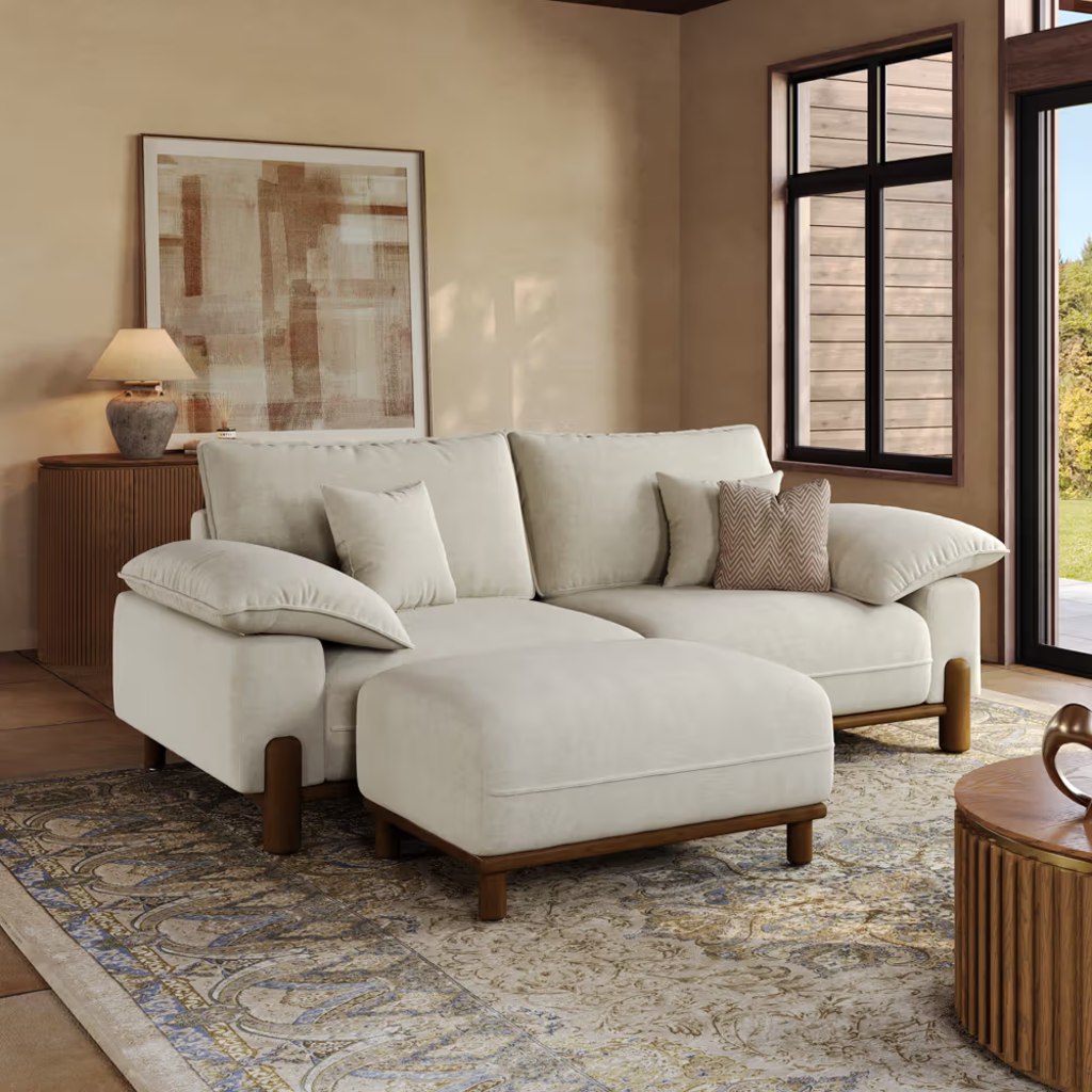 Modern living room with a white sofa, ottoman, patterned rug, wooden sideboard, and warm natural light from large windows.