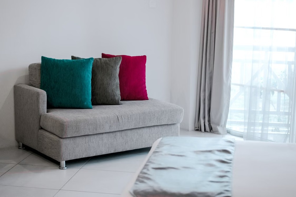 Compact gray sleeper chair with teal, charcoal, and magenta cushions beside a large window in a bright, small living room.