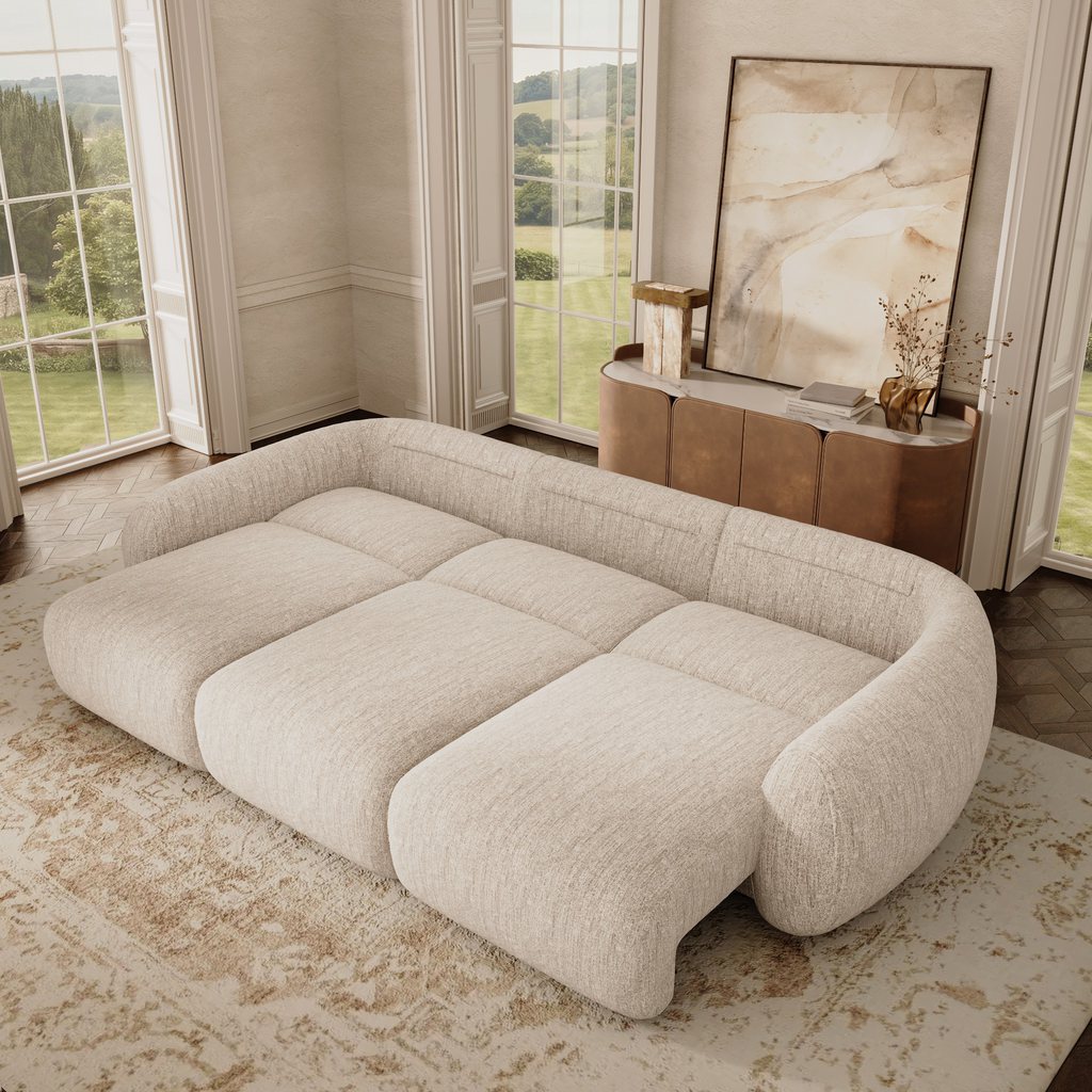 Overhead view of a beige three-seater power sofa bed fully extended into a spacious lounging surface in a bright living room with tall windows.