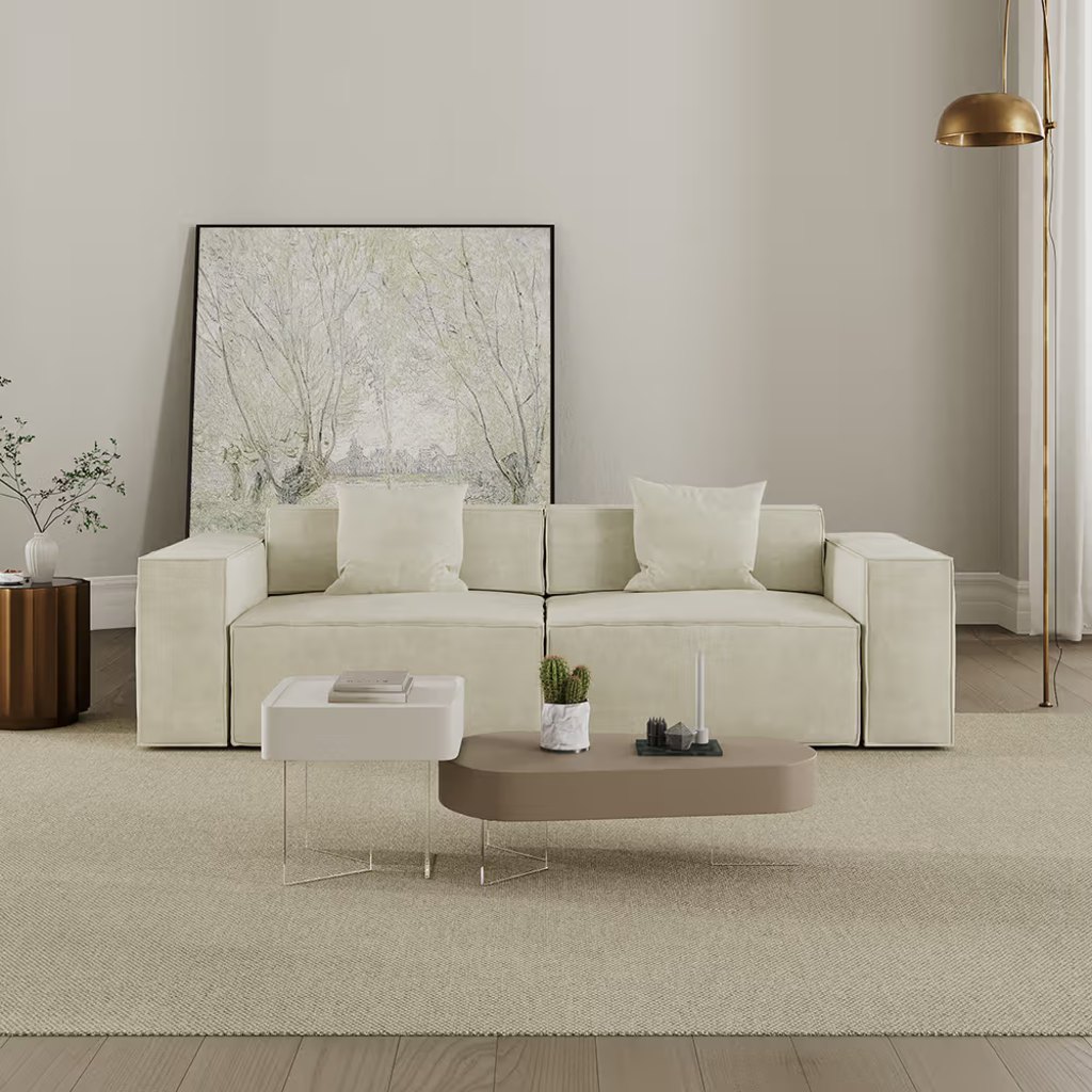 Cream-colored sleeper sofa with wide square arms and two white pillows in a minimalist living room with modern coffee tables and neutral decor.