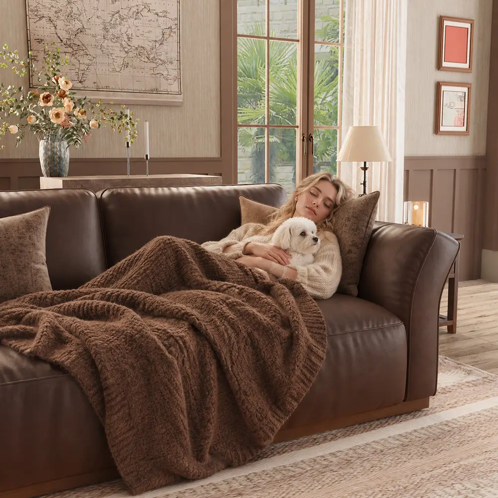 Woman cuddling a small white dog on a dark brown leather sofa under a soft brown blanket in a cozy, neutral-toned living room.