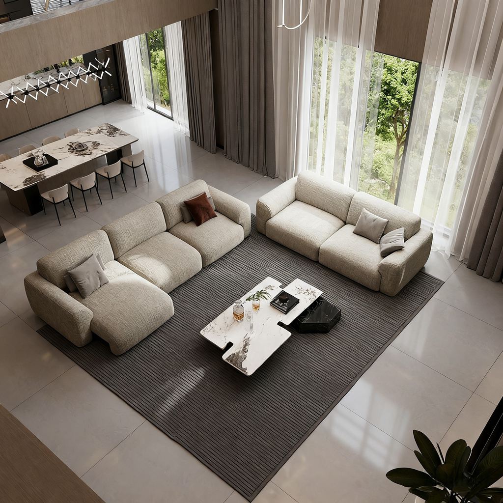 Spacious modern living room with two beige sofa beds, a marble coffee table, a large gray rug, and floor-to-ceiling windows overlooking greenery.