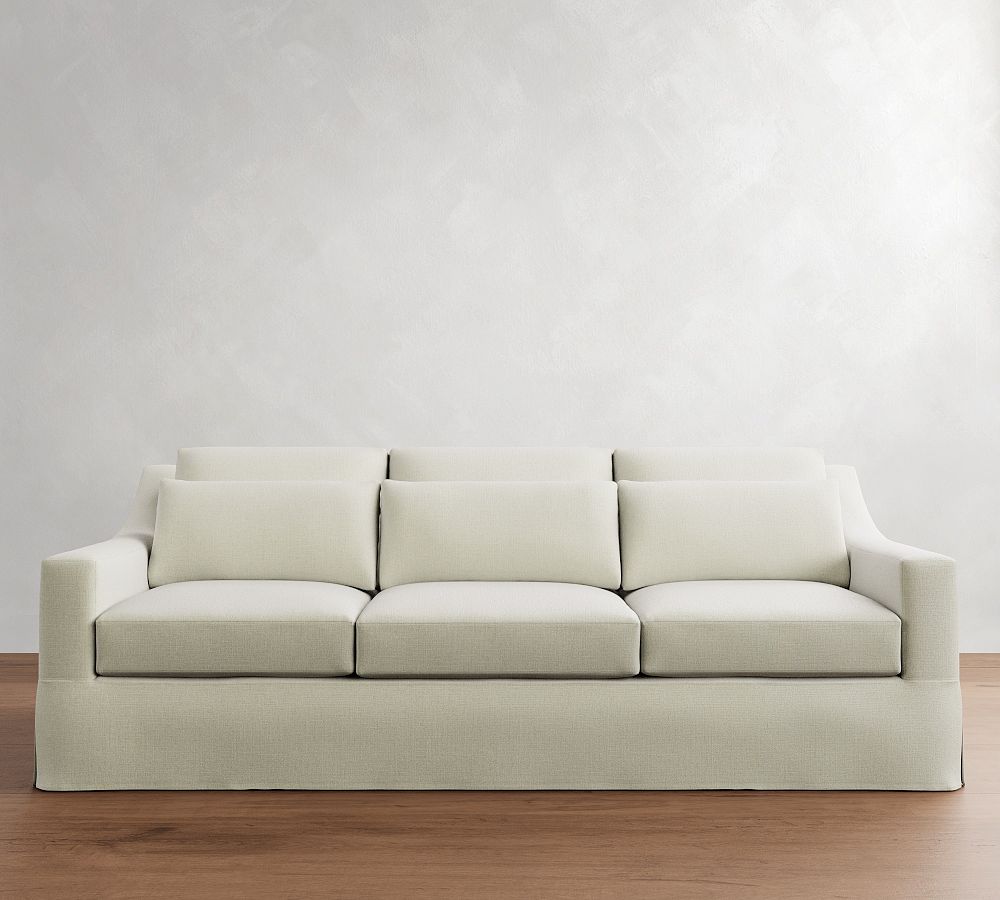 Cream-colored three-seat sofa with plush back cushions, wide armrests, and a tailored slipcover against a light gray wall.