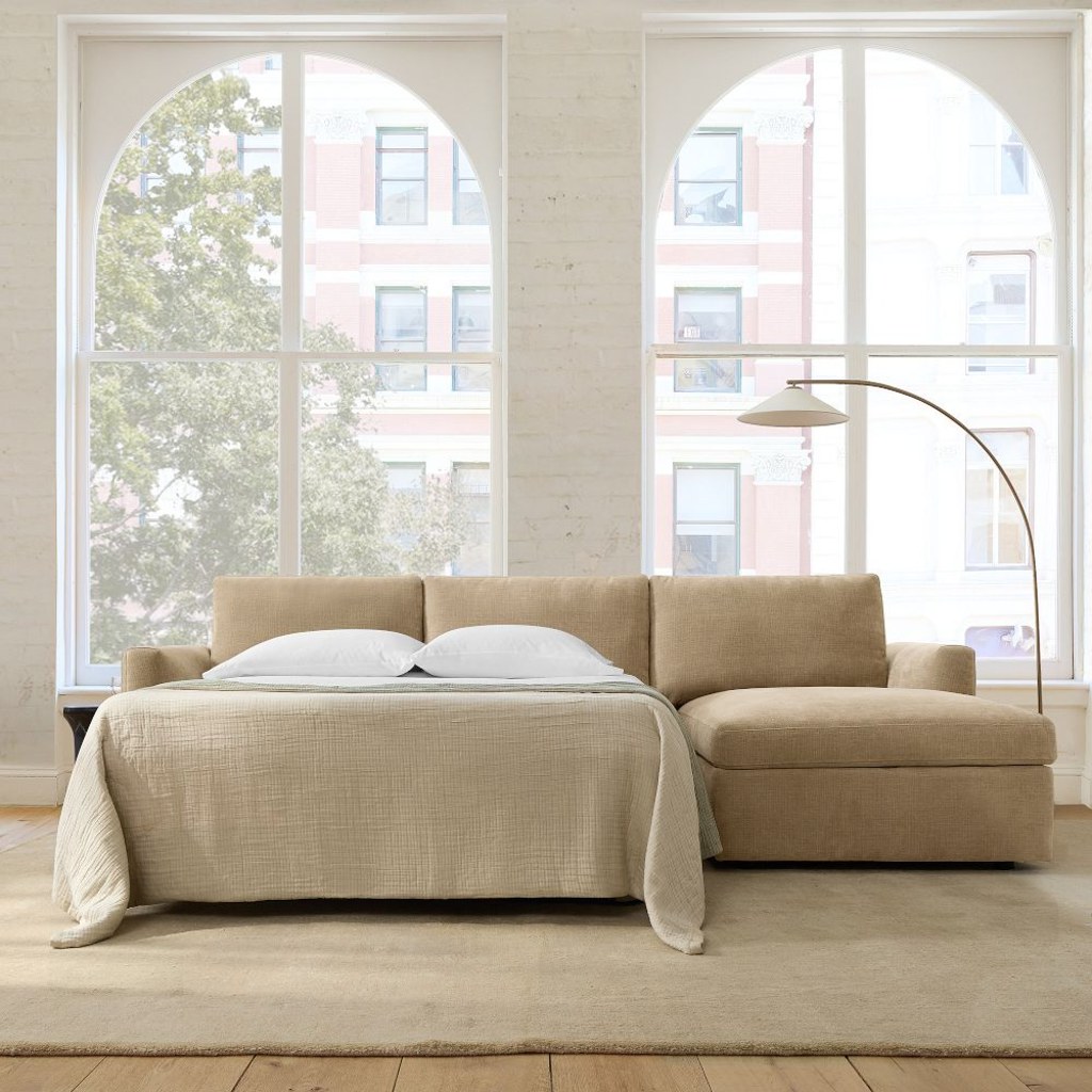 Beige sleeper sectional sofa with a pull-out bed and chaise lounge in a bright living room with large arched windows.