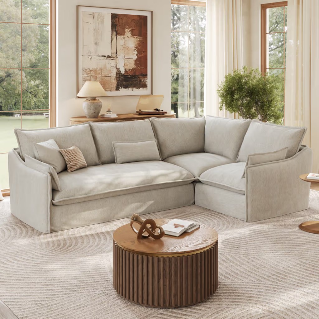Modern living room with a large cream sectional sofa, round wooden coffee table, abstract wall art, and floor-to-ceiling windows.